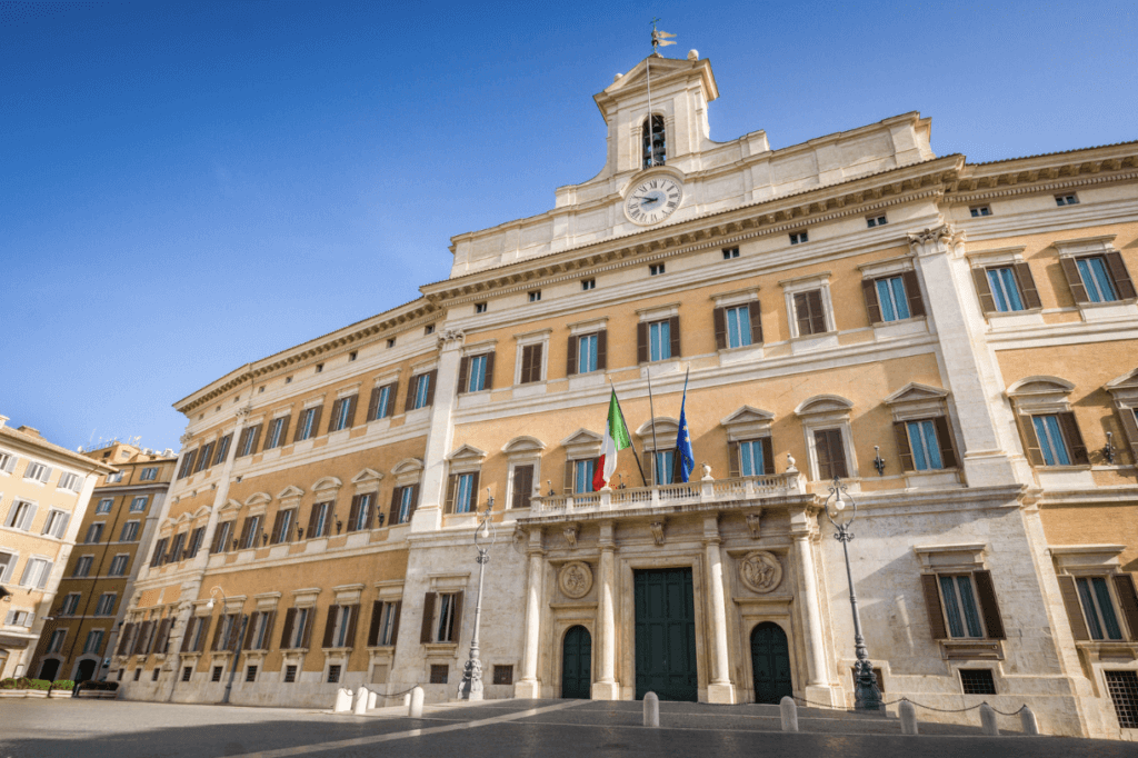 In primo piano, palazzo Montecitorio a Roma e la piazza antistante. Dietro, un cielo terso e la manovra di bilancio.