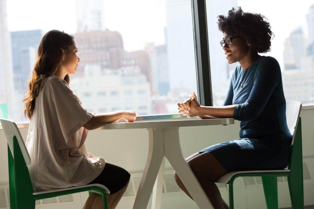 Due donne conversano sedute a un tavolo, sorridendo. Bonus psicologo.